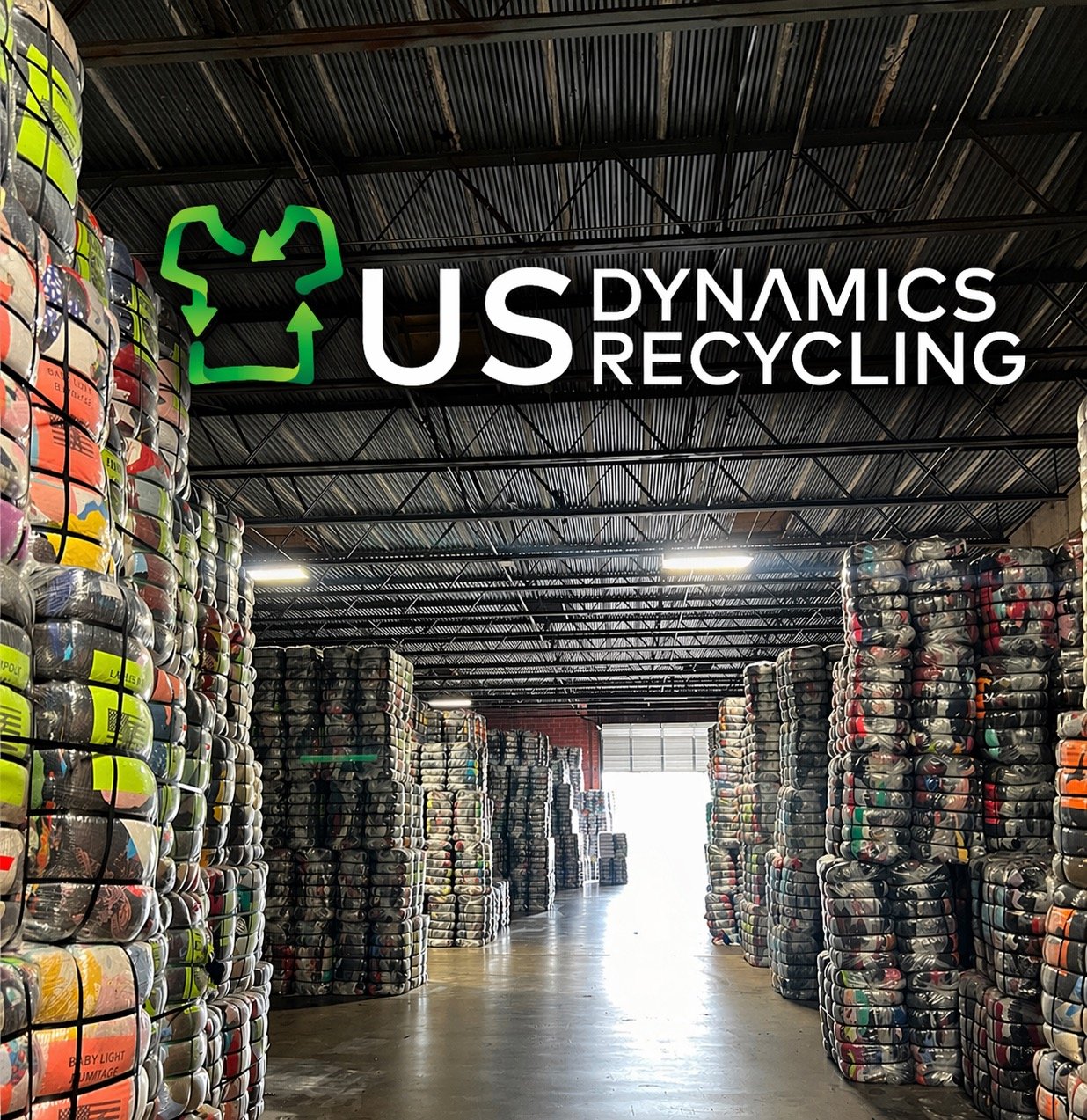 US Dynamics logo over a wall of used clothing bales